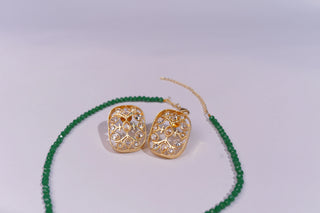 A pair of gold filigree stud earrings with clear stones, displayed next to a green beaded necklace on a light gray background.